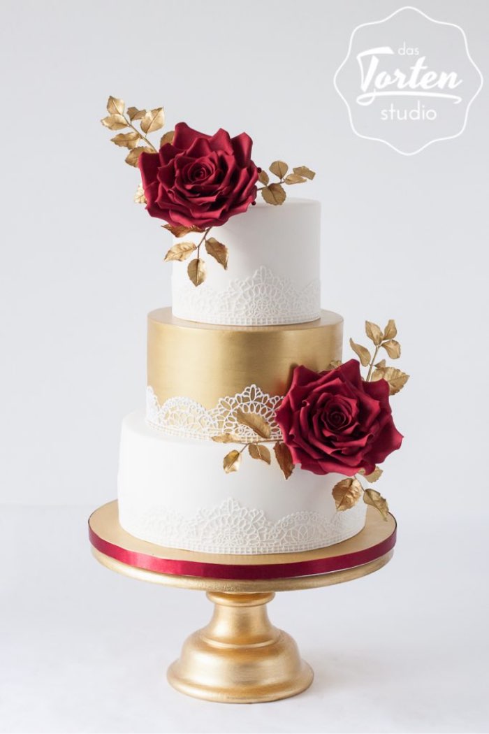 Dreistöckige Hochzeitstorte in Weiß und Gold, dekoriert mit weißer, essbarer Spitze, dunkelroten Zuckerrosen und goldenen Blättern