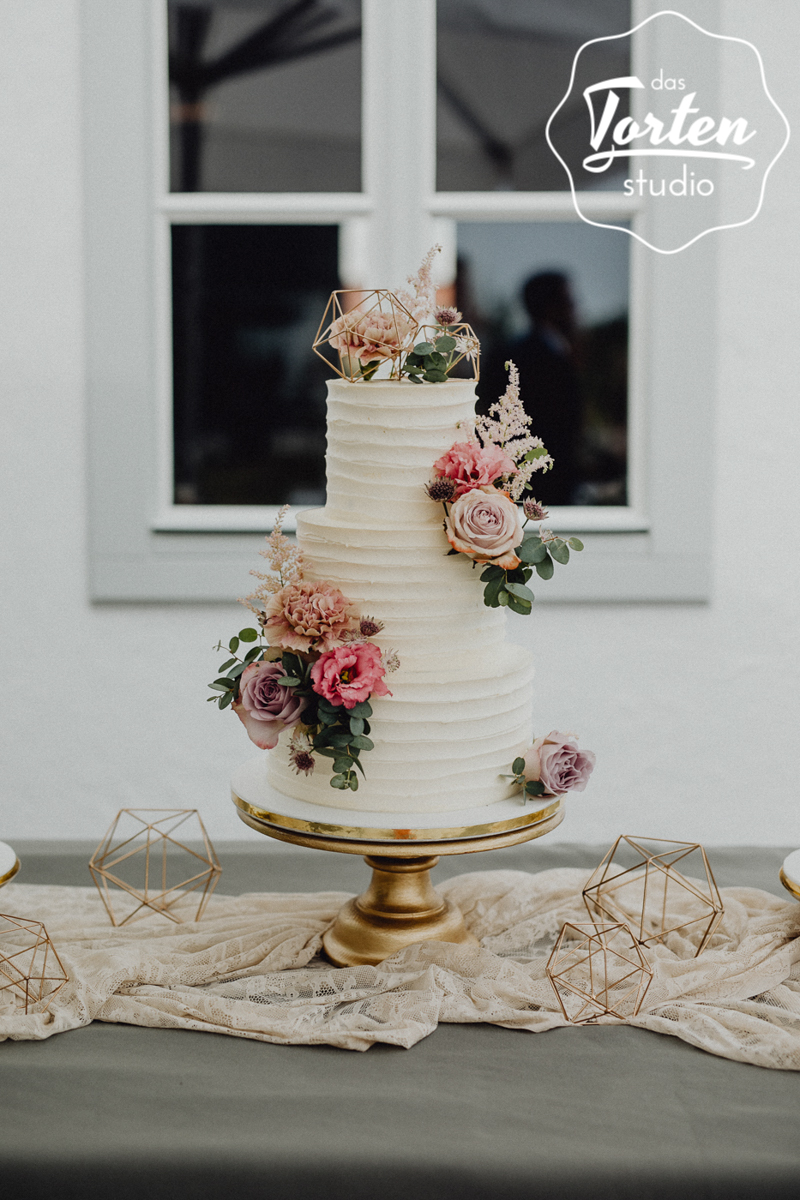 Dreistöckige Buttercreme-Torte mit Streifenoptik auf einem goldenen Tortenständer, dekoriert mit echten Blumen in pudrigen Rosa- & Pinktönen