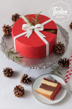Weihnachtstorte Punschtorte eingedeckt mit rotem Fondant und weißer Zuckerschleife, auf Tortenständer, Tortenstück auf Teller, Rosmarin und Bockerl als Deko